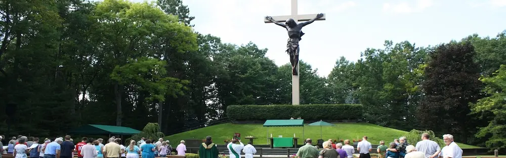 A large crucifix of Jesus Christ at the Cross in the Woods towers over a group of onlookers in a wooded outdoor area.
